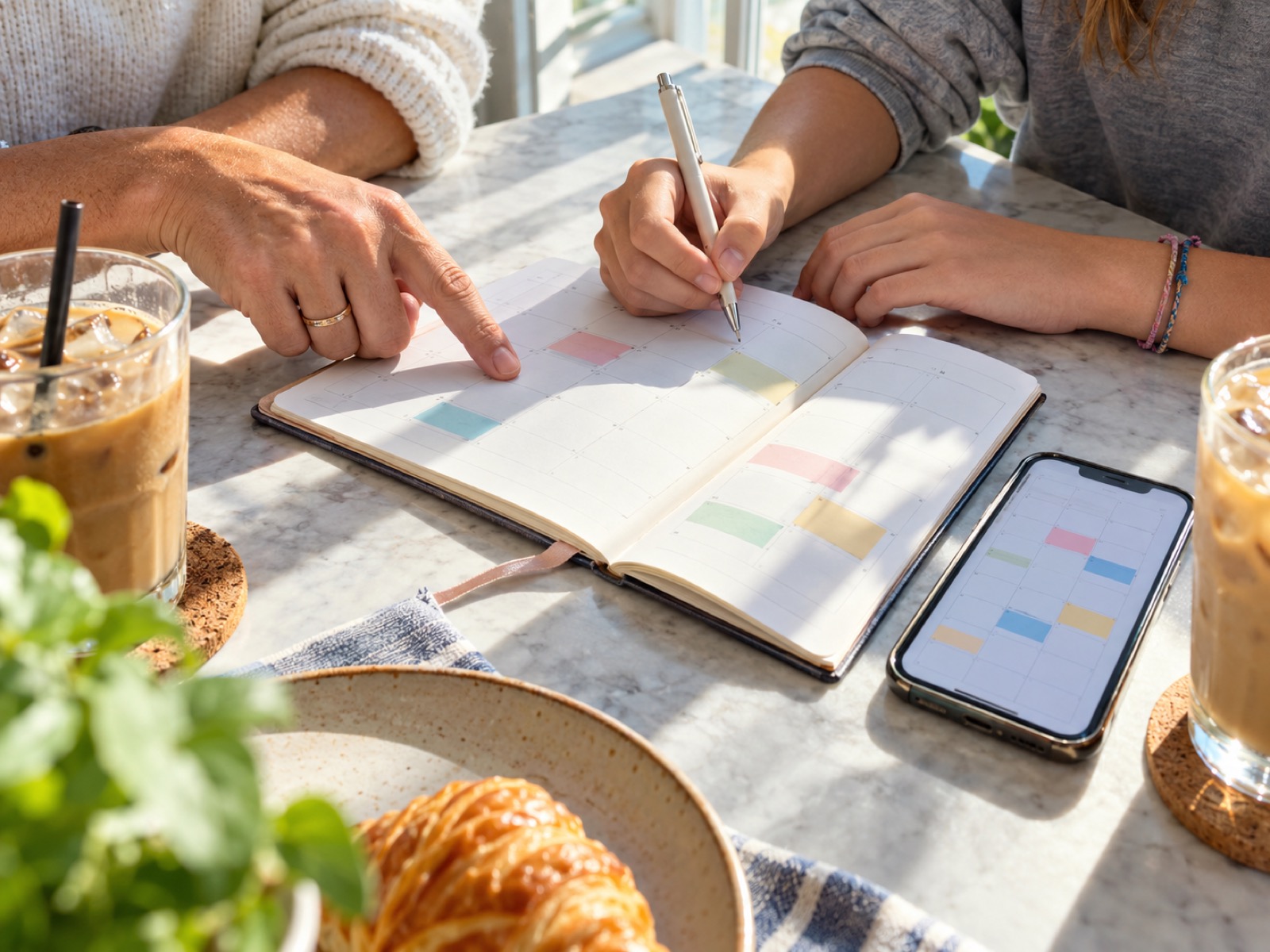 カフェのテーブルで、親子が手帳とスマートフォンのカレンダーに夏休みの予定を書き込む手元